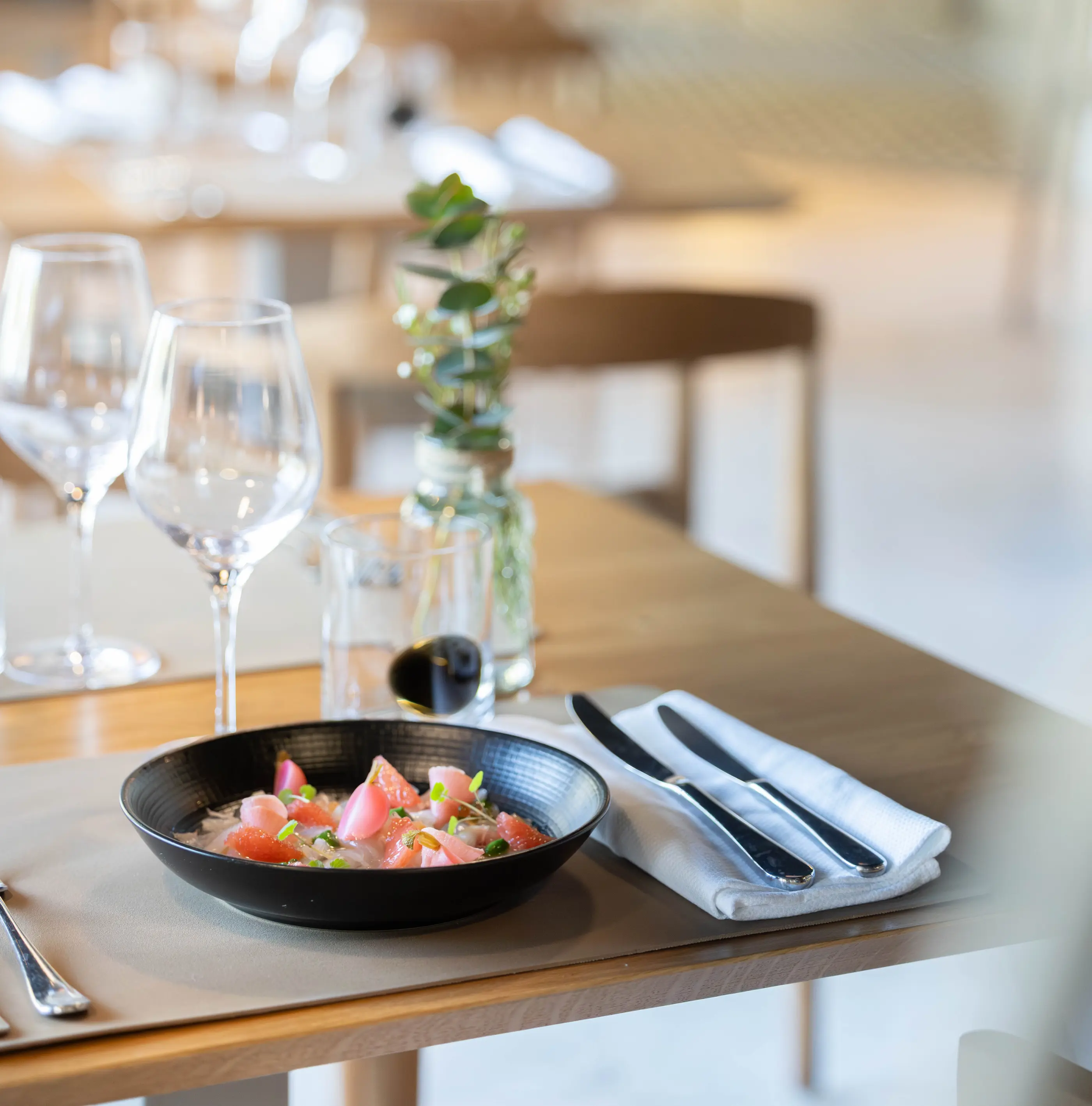 Un bol contenant une salade de tomates et des herbes, posé sur une table avec des couverts, un verre et une bouteille avec une plante verte