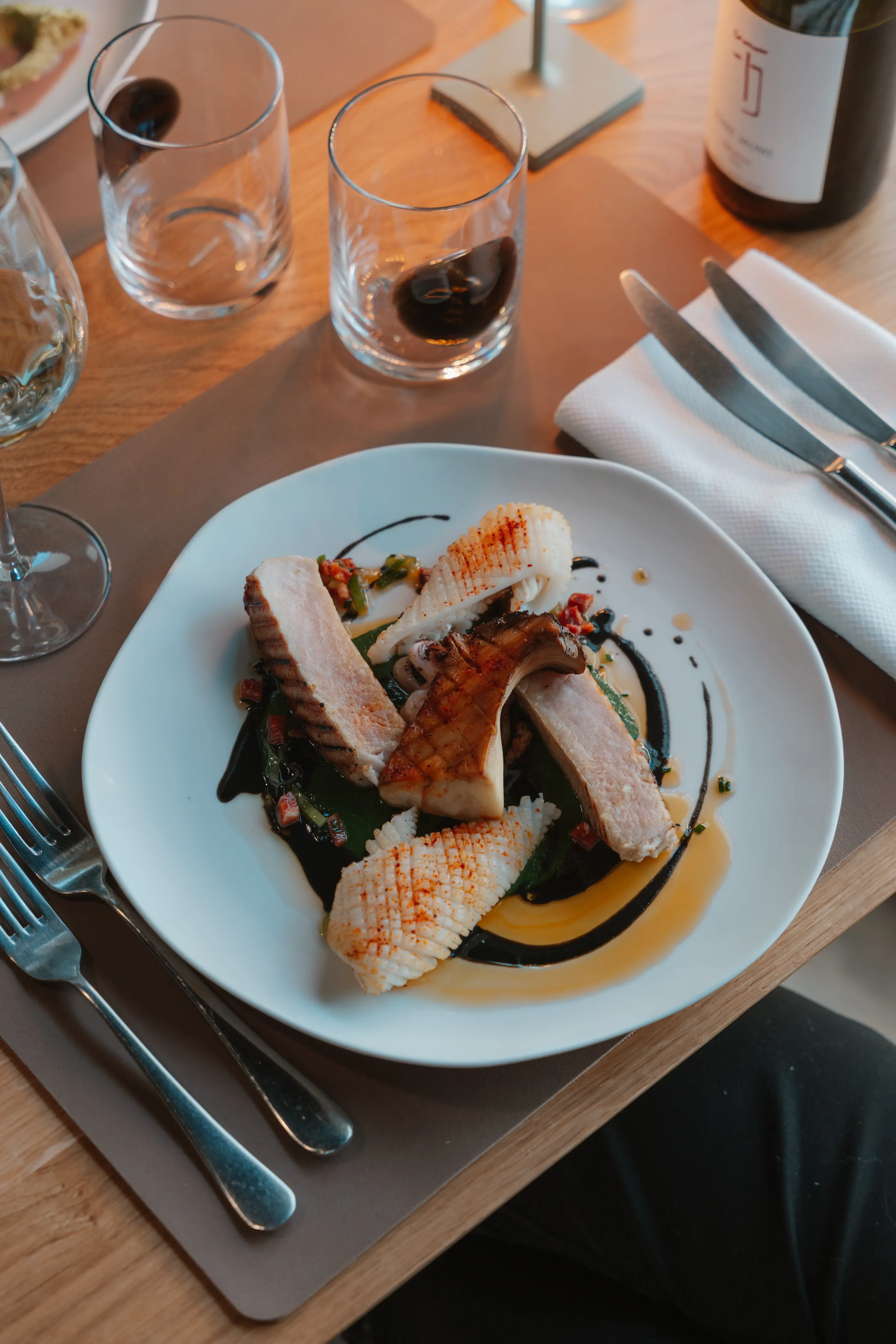Un plat de poisson grillé avec des légumes et une sauce, accompagné de couverts, posé sur une table avec des verres et une bouteille de vin