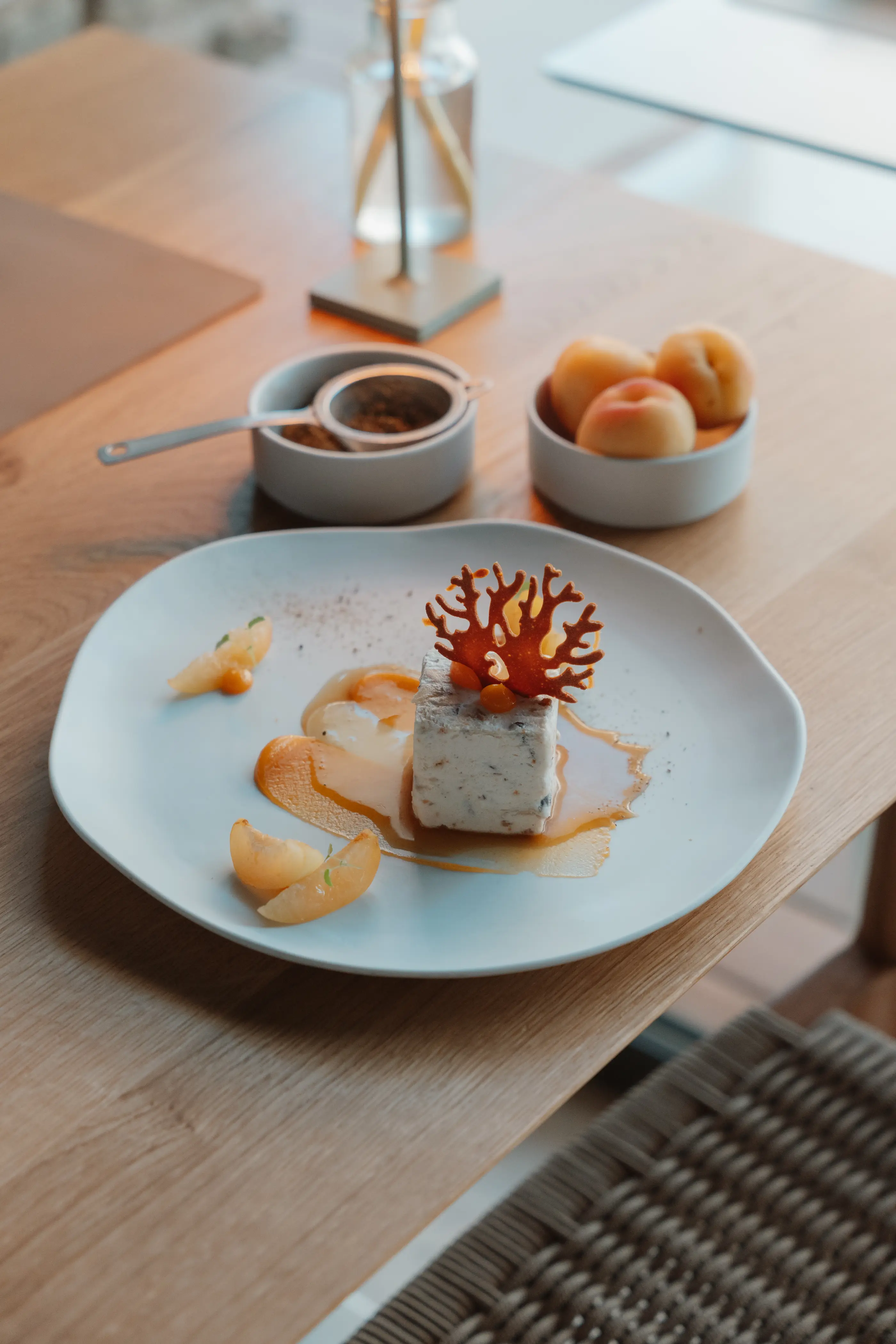 Un dessert à base de mousse blanche et de caramel décoré de zestes d'agrumes et d'une feuille de caramel rouge, accompagné de fruits et d'une sauce dans des petits bols sur une table en bois