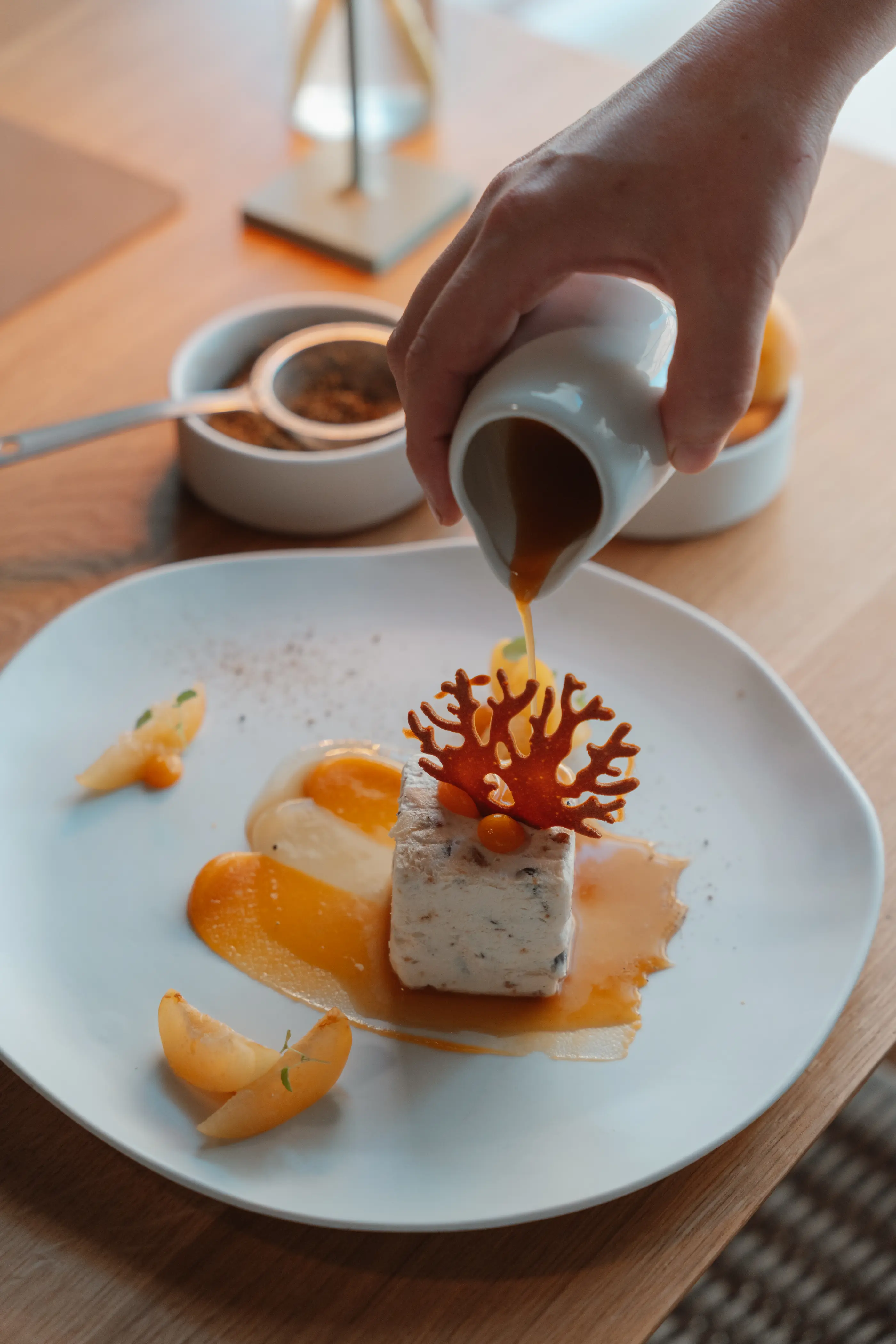 Une main verse une sauce sur une assiette contenant un dessert composé de fruits et d'un carré blanc décoré d'un motif en sucre