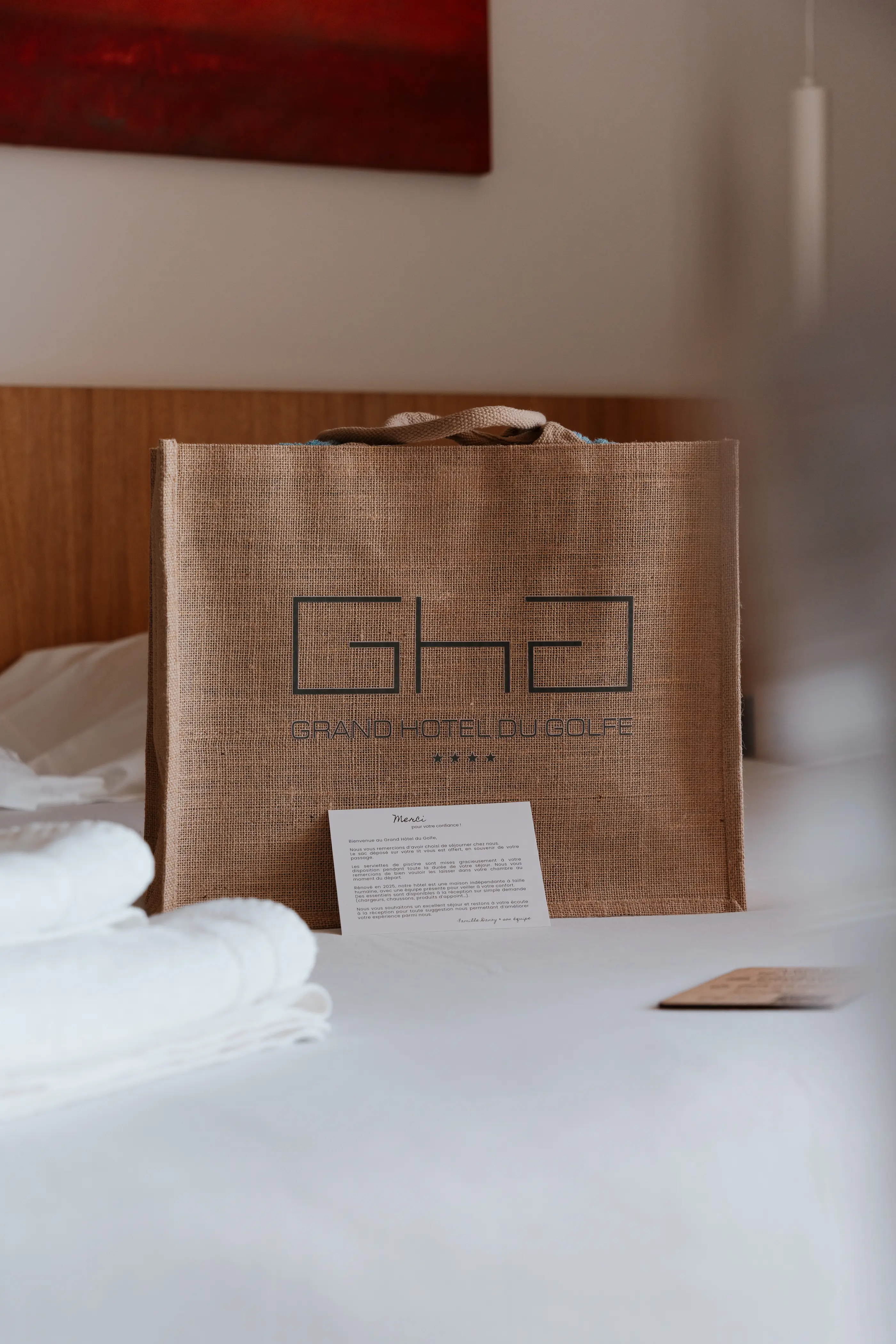 Un sac en jute avec le logo "GH" et le texte "GRAND HOTEL DU GOLFE" posé sur un lit avec une note manuscrite
