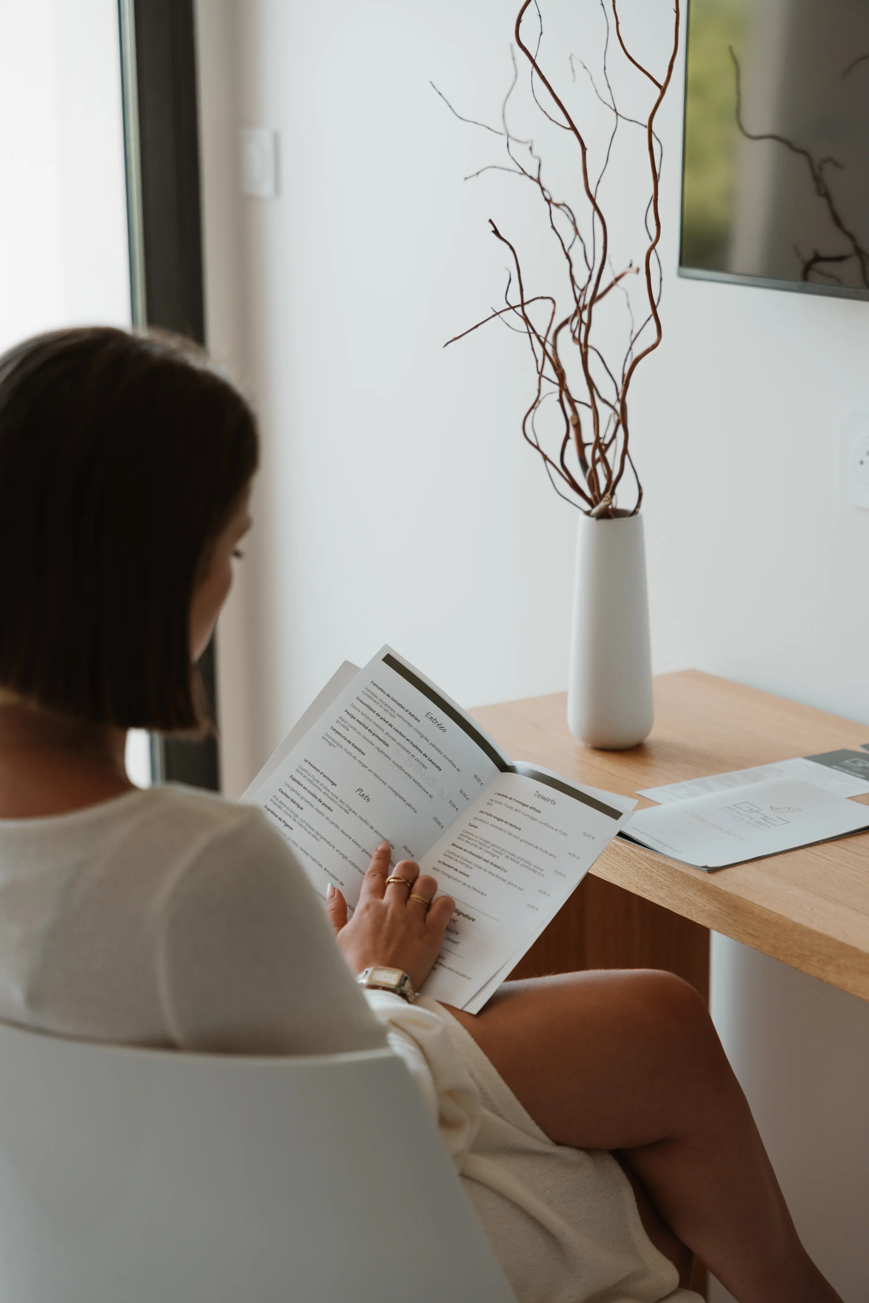 Une personne assise lit un document sur une table avec un vase contenant des branches séchées