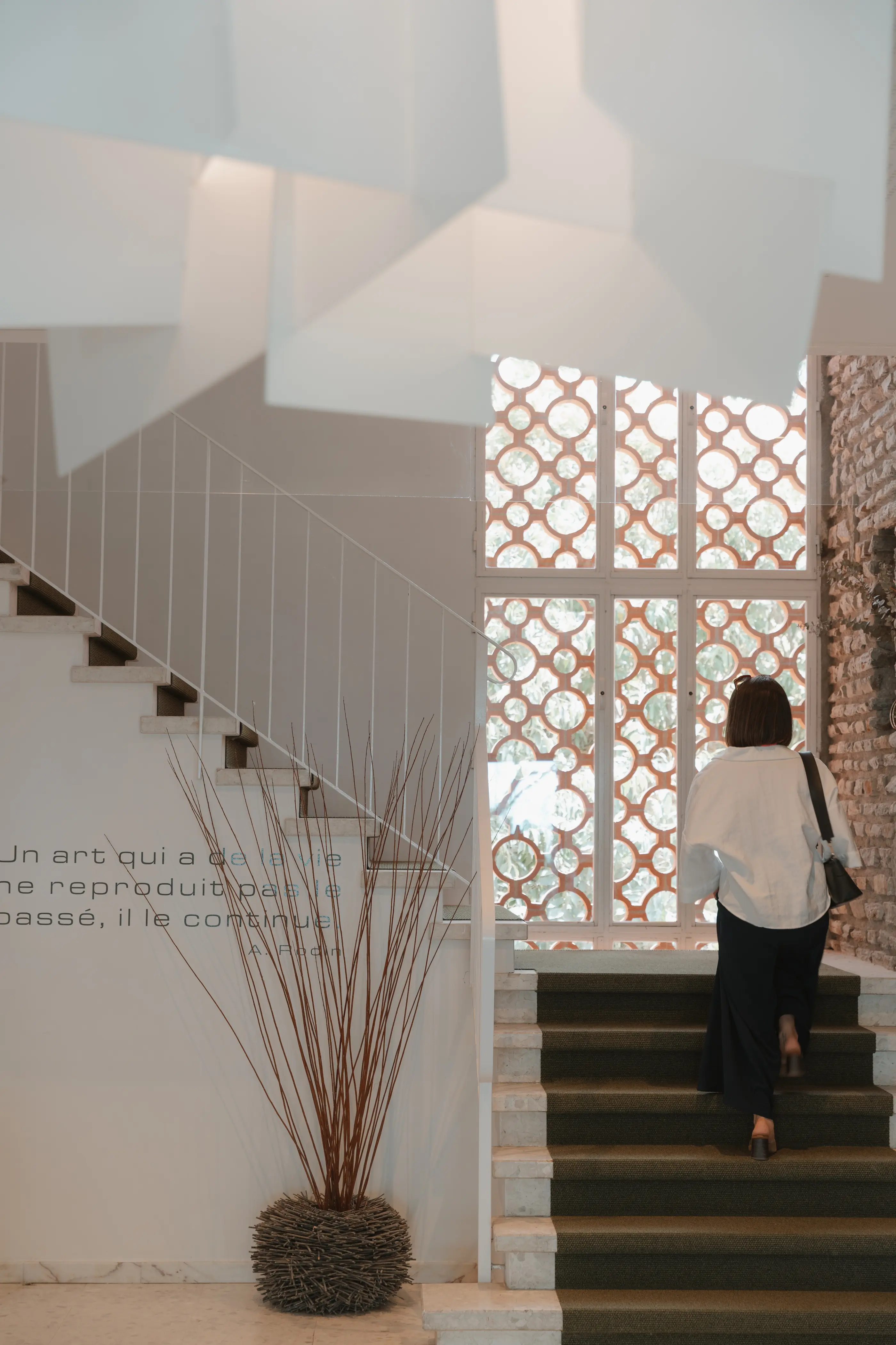 Une personne monte un escalier près d'une fenêtre à motifs circulaires, avec une plante en pot à gauche