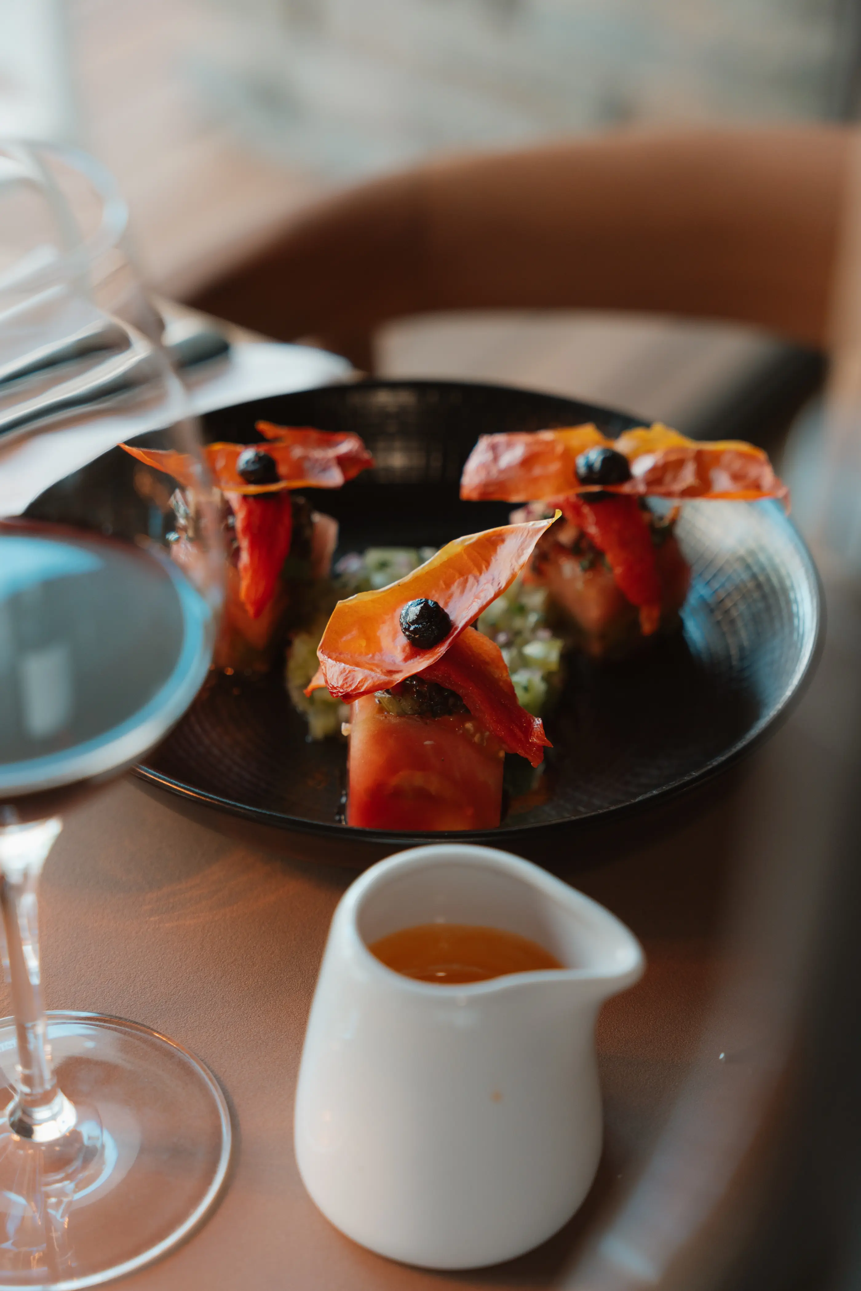 Un plat avec des morceaux de tomates et de bacon décorés de baies, accompagné d'une petite saucière blanche, posé sur une table avec un verre à vin