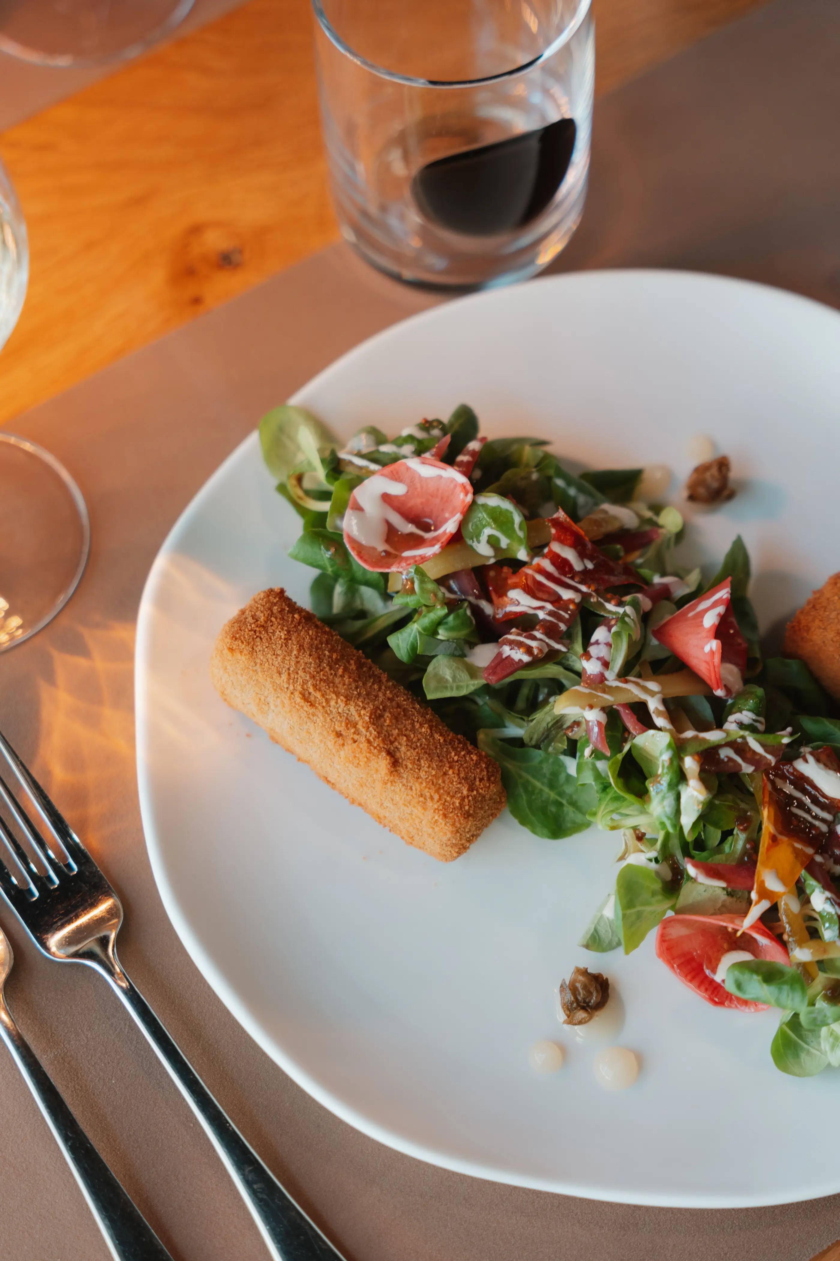 Une assiette contenant une salade verte avec des tomates, des noix, des copeaux de parmesan et une croquette, accompagnée d'un verre de vin rouge et d'ustensiles